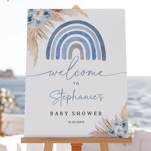 May include: A white baby shower welcome sign with a watercolor rainbow in shades of blue. The sign includes the text "welcome to Stephanie's BABY SHOWER" and is decorated with floral accents and pampas grass.