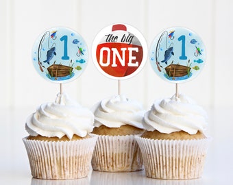 Puede incluir: Tres cupcakes con glaseado blanco y un adorno de cumpleaños con temática de pesca. Los adornos presentan una caña de pescar azul y blanca, un flotador de pesca rojo y blanco con el texto "the big ONE", y un barco de pesca azul y blanco con un pez y el número "1".