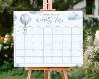 Könnte beinhalten: Ein weißer Kalender mit dem Text "GUESS BABY LOGAN'S birthday date" in blauer Schrift. Er zeigt eine blau-weiße Heißluftballon-Grafik und blaue Aquarellwolken. Der Kalender wird auf einer Holzstaffelei angezeigt.