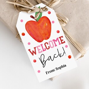 Può includere: Un'etichetta regalo con un'illustrazione ad acquerello di una mela e le parole "WELCOME Back!" in rosso e rosa. L'etichetta è decorata con pois rossi e rosa e il nome "Sophia". L'etichetta è legata a un regalo confezionato con rafia.