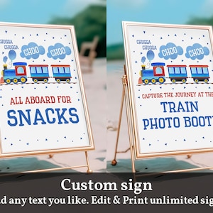 May include: Two gold-colored easel stands with white signs. The sign on the left says "All aboard for snacks" with a cartoon train and the sign on the right says "Capture the journey at the train photo booth" with a cartoon train.