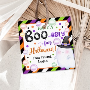 May include: A Halloween-themed gift tag with a cute ghost wearing a witch hat. The tag has a black and white striped background with green and purple accents. The text on the tag reads "Have a Boo-bbly fun Halloween! Your friend, Logan".
