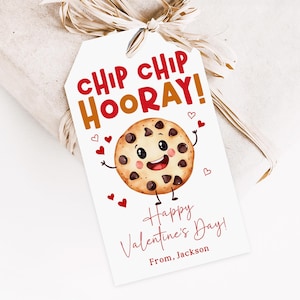 May include: A gift tag with a cartoon cookie illustration. The tag reads "CHIP CHIP HOORAY!" in red and orange lettering. Below the cookie, it says "Happy Valentine's Day!" and "From, Jackson" in red script. The tag is tied to a wrapped gift.