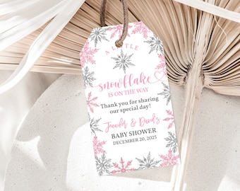 以下が含まれることがあります： ピンクの雪の結晶のデザインと「A Little Snowflake is on the Way」と「Jennifer & David's Baby Shower December 20, 2025」のテキストが書かれた白いタグ。