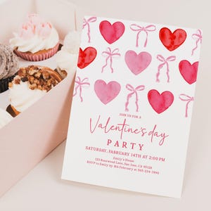 Puede incluir: Una invitaci&oacute;n a una fiesta de San Valent&iacute;n con corazones y lazos de acuarela. La invitaci&oacute;n est&aacute; sobre un fondo blanco e incluye los detalles de la fiesta. Una caja de cupcakes con varios ingredientes est&aacute; en el fondo.