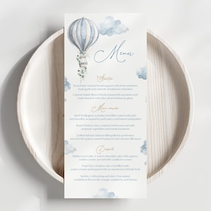 May include: A menu card with a watercolor illustration of a blue and white hot air balloon and clouds. The menu includes starters, main courses, and desserts. The card is placed on a light wooden plate.