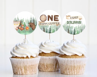 May include: Three cupcakes with white frosting and decorative toppers. The toppers feature watercolor illustrations of a camping theme, including a tent, campfire, fox, and bear. One topper says "ONE HAPPY CAMPER", and another says "CAMP BENJAMIN".