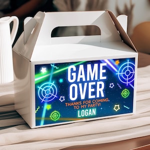 May include: White party favour box with a handle, featuring a blue and green graphic that reads "GAME OVER". The design includes neon-style targets, stars, and the name "LOGAN". The text "THANKS FOR COMING TO MY PARTY!" is also included.