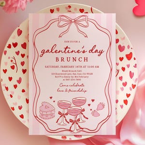 May include: A pink invitation for a Galentine's Day brunch, featuring a striped background and a decorative frame. The invitation includes the date, time, location, and RSVP information. The invitation is on a heart-decorated plate.
