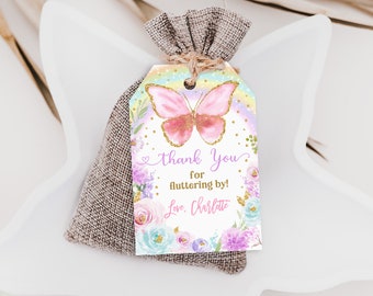 May include: A pink and gold butterfly with glitter accents on a white tag with a rainbow background. The tag has the text "Thank You for fluttering by! Love, Charlotte" and is decorated with purple, pink, and blue flowers.