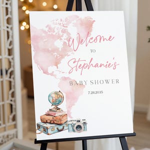 Puede incluir: Un letrero blanco sobre un caballete negro con el texto "Welcome to Stephanie's Baby Shower" en escritura rosa. Un mapa del mundo en acuarela está en el fondo. El letrero incluye un globo terráqueo, libros apilados, maletas vintage y una cámara.