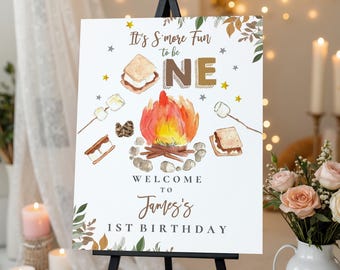 Puede incluir: Un letrero blanco con una ilustración de acuarela de una fogata, s'mores y el texto "It's S'more Fun to be ONE". El letrero da la bienvenida a los invitados al 1er cumpleaños de James.