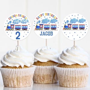 May include: Three cupcake toppers featuring a blue train theme. The toppers depict a train with red, blue and yellow details. One topper reads "Happy Birthday Jacob", another says "Chugga Chugga Two Two 2", and the third topper shows a train with blue and white clouds.