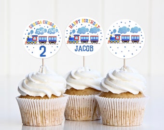 May include: Three cupcake toppers with a blue train theme. The toppers feature a train with red, blue, and yellow details. One topper says "Happy Birthday Jacob", another says "Chugga Chugga Two Two 2", and the third topper has a train with blue and white clouds.