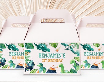 May include: Three white favor boxes with a dinosaur theme. The boxes have a handle and are decorated with green leaves, orange volcanoes, and blue and green dinosaurs. The text on the boxes reads "Benjamin's 1st Birthday".