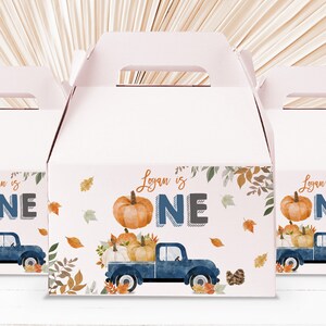 Può includere: Un set di tre scatole regalo blu e bianche a tema autunnale. Le scatole presentano un camioncino blu con zucche e foglie autunnali. Il testo sulle scatole recita "Logan is One".