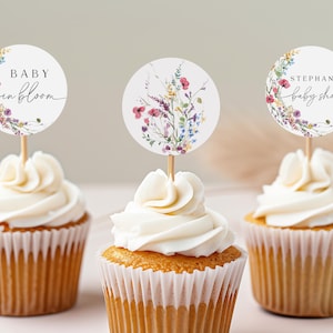 May include: Three cupcakes with white frosting topped with round floral themed paper cupcake toppers. The toppers say "Baby in Bloom", "Stephanie's Baby Shower", and feature a watercolour floral design.