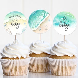 May include: Three cupcakes with white frosting topped with round paper cupcake toppers. The toppers feature a watercolour design of ocean waves and a sandy beach. The text on the toppers reads "Welcome baby", "Oh baby!" and a beach scene.