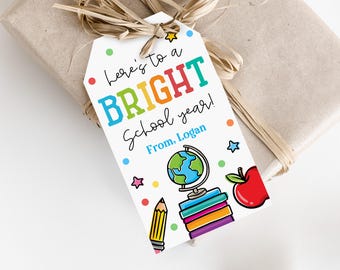 May include: A gift tag with the text "Here's to a BRIGHT School year!" in colorful letters. The tag features a globe, books, a pencil, and an apple illustration. The gift is wrapped in brown paper and tied with raffia.