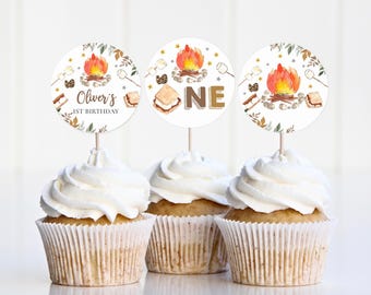 Puede incluir: Tres cupcakes con glaseado blanco, cada uno coronado con un adorno redondo. Los adornos presentan un diseño de fogata y el texto "Oliver's 1st Birthday" y "ONE". Los cupcakes están en moldes de papel.