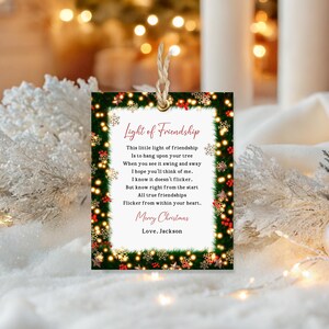 May include: A rectangular Christmas gift tag with a green border adorned with red berries, snowflakes, and lights. The tag features the text "Light of Friendship" and a poem, with "Merry Christmas, Love, Jackson" at the bottom. A twine string is attached.