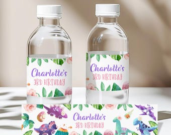 Può includere: Due bottiglie d'acqua in plastica trasparente con tappi bianchi ed etichette personalizzate con illustrazioni ad acquerello di dinosauri, foglie tropicali e fiori rosa. Le etichette riportano il testo Charlotte's 3rd BIRTHDAY.