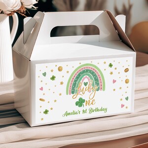 May include: White favor box with a green shamrock and rainbow design. The box has gold glitter accents and the text "Lucky One" and "Amelia's 1st Birthday".