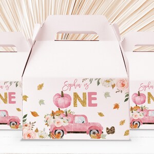 Può includere: Tre scatole rosa per bomboniere a tema autunnale. Ogni scatola presenta un camion rosa che trasporta zucche e fiori, con la scritta "Sophia is ONE" in glitter dorati. Foglie autunnali e accenti floreali completano il design.