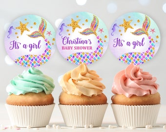 May include: Three cupcakes decorated with mermaid-themed baby shower toppers. The toppers are round and feature a mermaid tail, a starfish, and a seahorse. The text on the toppers reads "It's a girl" and "Christina's Baby Shower".