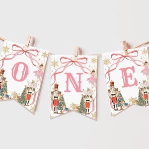May include: A decorative banner spelling "ONE" for a first birthday. Each pennant features a pink letter, a nutcracker, a ballerina, and a mouse. The banner is decorated with gold accents and a pink ribbon.