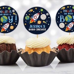 May include: Three cupcakes decorated with blue and white space-themed cupcake toppers. The toppers feature a rocket ship, planets, stars, and the text "Houston, we have a boy!" and "Jessica's Baby Shower".