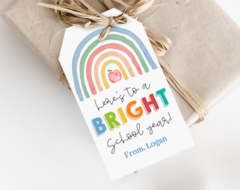 Könnte beinhalten: Ein Geschenkanhänger mit einem Aquarell-Regenbogen-Design und dem Text "Here's to a BRIGHT School year!" in bunten Buchstaben. Der Anhänger enthält den Namen "From, Logan" und ist an einem verpackten Geschenk mit Bastband befestigt.