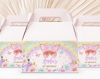 May include: Three pink and white favor boxes with a rainbow and butterfly design. The boxes have a handle and are decorated with gold glitter. The text on the boxes reads "Amelia's 1st Birthday".
