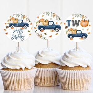 May include: Three cupcake toppers featuring a blue truck carrying pumpkins and autumn leaves. The toppers read "Happy Birthday", "Two", and depict a truck with pumpkins.
