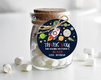 May include: A small glass jar filled with white marshmallows with a round, blue and black thank you tag attached. The tag features a rocket ship, planets, and stars. The text on the tag reads "Thank you for making my party a blast! Love, Nathan."