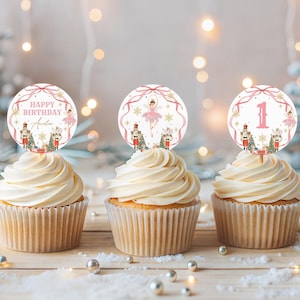May include: Three cupcakes with white frosting, each topped with a round, pink and gold birthday cake topper. The toppers feature a ballerina, nutcracker, and the text "Happy Birthday Amelia" or the number "1". The background includes Christmas decorations.