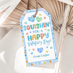 May include: A Valentine's Day gift tag with a blue border and colorful polka dots. The tag reads "Squishin' You a Happy Valentine's Day!" with hearts and the name "Logan" at the bottom. A brown string is attached.
