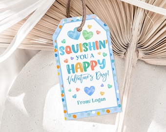 Pode incluir: Uma etiqueta de presente de Dia dos Namorados com uma borda azul e bolinhas coloridas. A etiqueta diz "Squishin' You a Happy Valentine's Day!" com corações e o nome "Logan" na parte inferior. Uma corda marrom está presa.