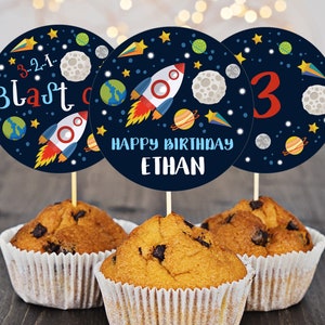 May include: Three round blue cupcake toppers with a space theme. The toppers feature a rocket, planets, stars, and the text "3...2...1... Blast Off" and "Happy Birthday Ethan".