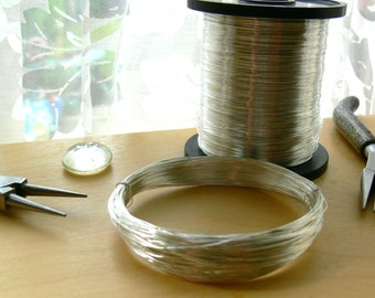 May include: A spool of silver wire with a coil of wire on a wooden surface. There are also a pair of pliers and a small glass bead.