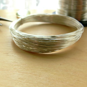 May include: A close-up of a silver wire coil, wound into a circular shape. The wire is shiny and reflective, and the coil is lying on a light brown wooden surface.