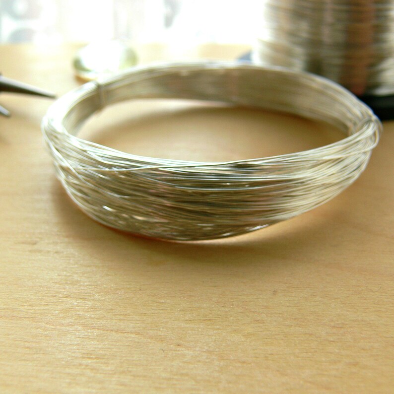 May include: A close-up of a silver wire coil, wound into a circular shape. The wire is shiny and reflective, and the coil is lying on a light brown wooden surface.