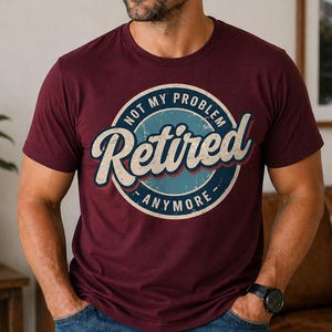 Op de afbeelding: Bordeauxrode crewneck t-shirt met een cirkelvormige afbeelding. De afbeelding toont het woord "Retired" in een vintage stijl, met de woorden "Not My Problem" en "Anymore" in wit, blauw en rood.
