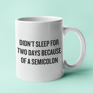 Op de afbeelding: Een witte keramische koffiemok met zwarte tekst die "Ik heb twee dagen niet geslapen vanwege een puntkomma." zegt.