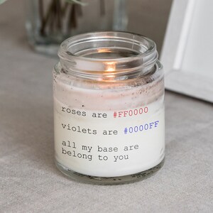以下が含まれることがあります： 白いラベルに「roses are #FF0000 violets are #0000FF all my base are belong to you」と書かれたガラス製のキャンドルジャー。キャンドルに火が灯され、ジャーは薄い茶色の表面の上に置かれている。