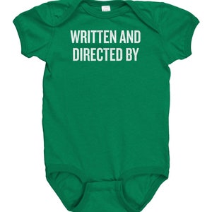 May include: A green baby onesie with the text "WRITTEN AND DIRECTED BY" in white block letters. The onesie is short-sleeved and has a simple, classic design. The garment is made of a soft material.