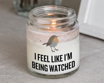 May include: A glass candle jar with a white label that says "I feel like I'm being watched". A small robin illustration is on the label. The candle is lit and the flame is visible.