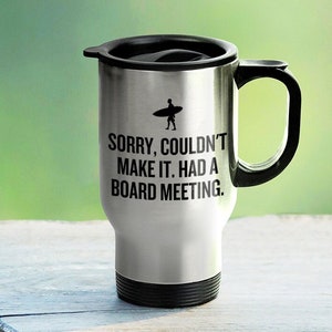 May include: A silver travel mug with a black lid and handle. The mug has a black silhouette of a surfer on a surfboard and the text "SORRY, COULDN'T MAKE IT. HAD A BOARD MEETING."