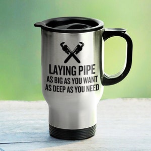May include: A silver travel mug with a black lid and handle. The mug has a black graphic of two crossed wrenches and the text "Laying Pipe As Big As You Want As Deep As You Need".