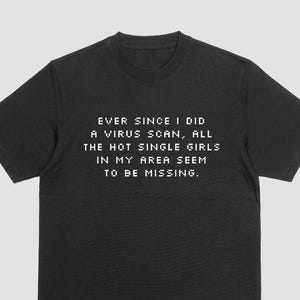May include: Black t-shirt with white text that reads: "EVER SINCE I DID A VIRUS SCAN, ALL THE HOT SINGLE GIRLS IN MY AREA SEEM TO BE MISSING." The shirt has short sleeves and a crew neck.